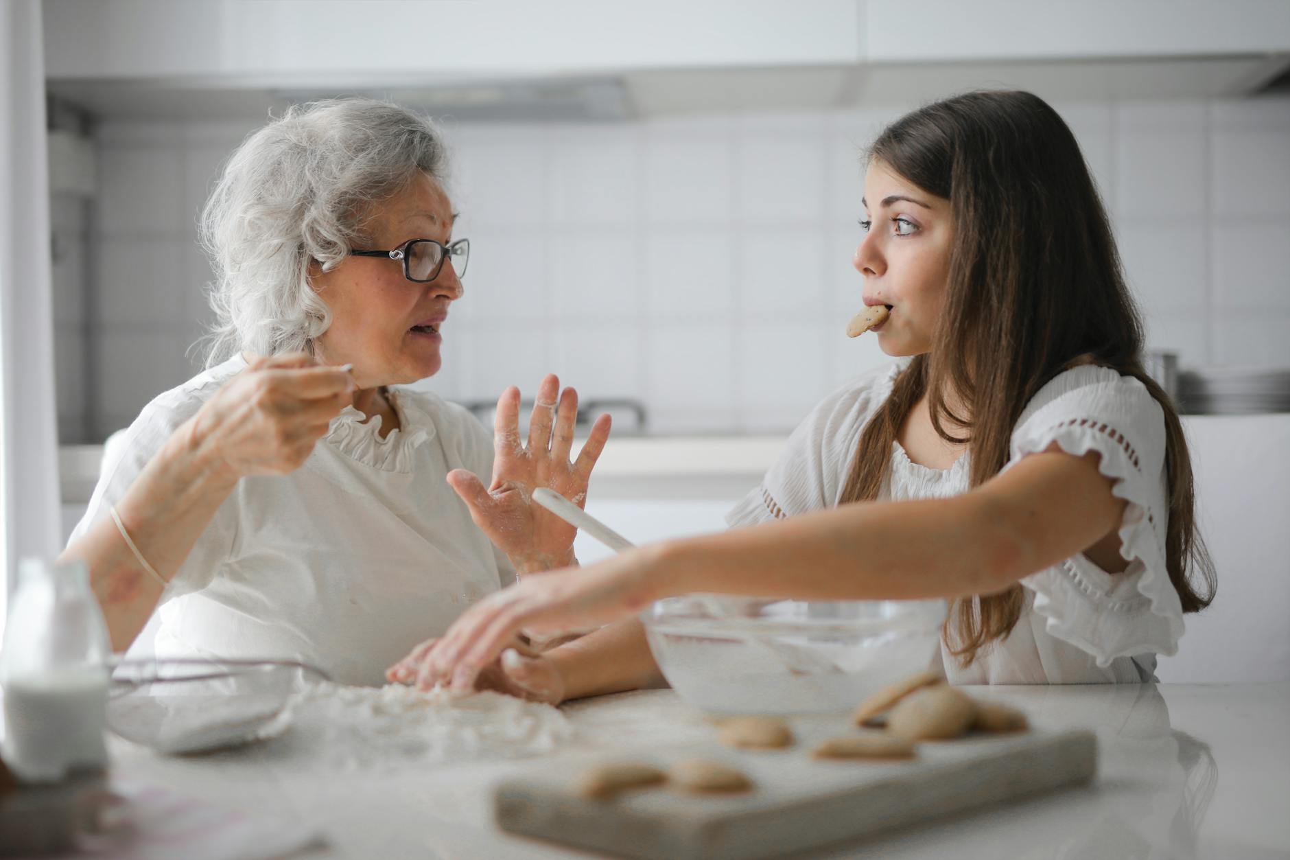 Pensive grandmother with granddaughter having interesting conversation while cooking together in light modern kitchen