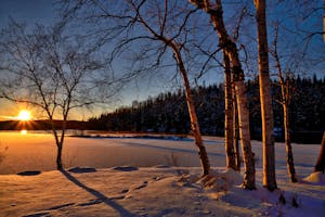 A serene winter sunset casting shadows on a frozen lake surrounded by snow and trees.