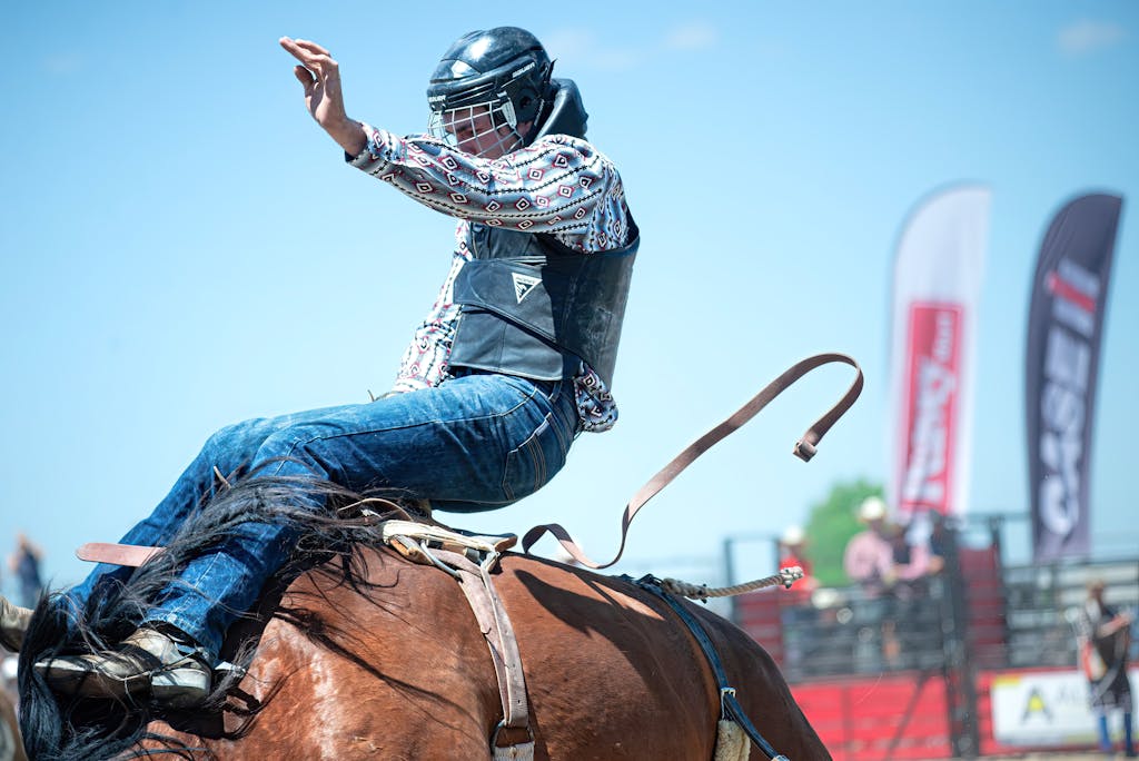 A cowboy in full gear riding a bucking horse at a rodeo event on a sunny day.