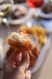 Delicious crispy battered fish piece held by hand with blurred chips in background.