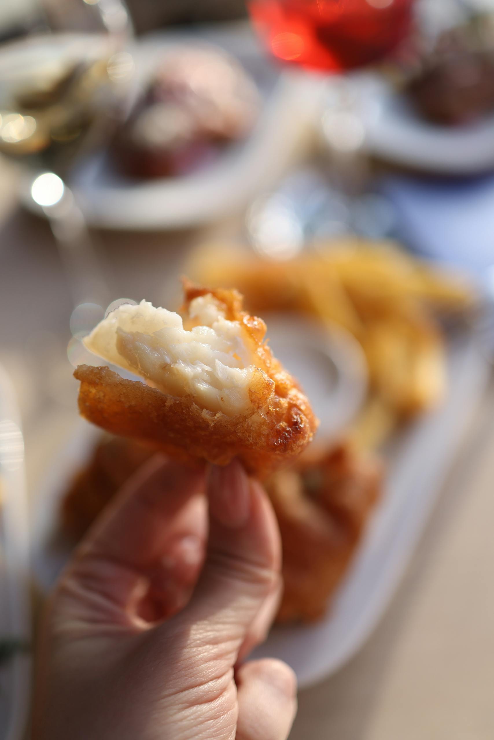 Delicious crispy battered fish piece held by hand with blurred chips in background.